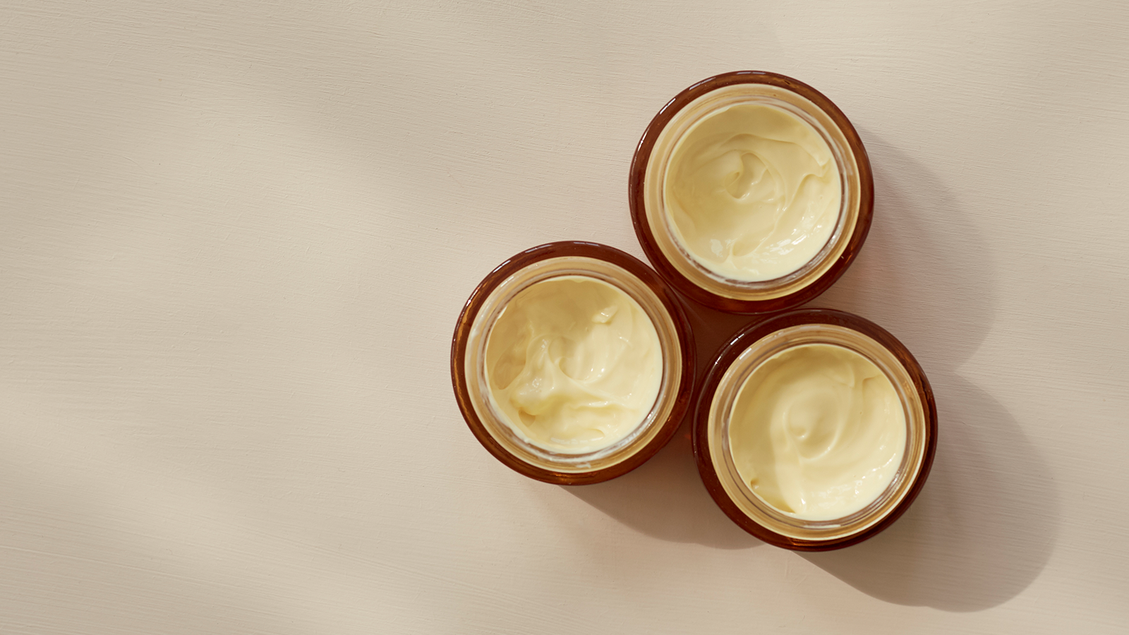 Tre åbne krukker med lys creme set ovenfra på en lys baggrund.