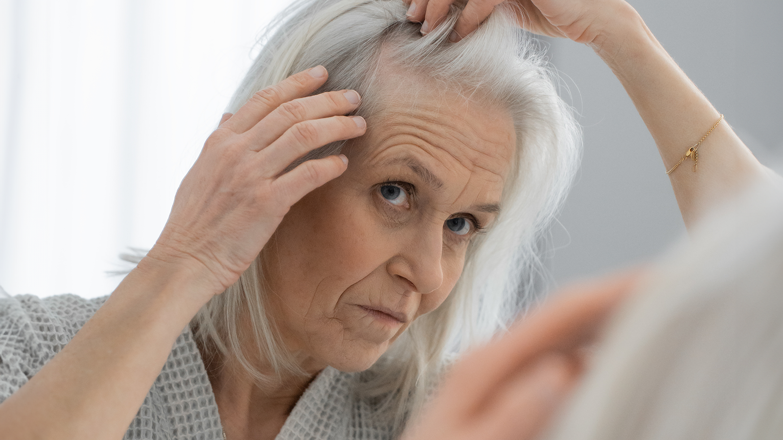 Hair Volume™ 17 Ældre kvinde ser bekymret på sit hår og undersøger hovedbunden foran et spejl.