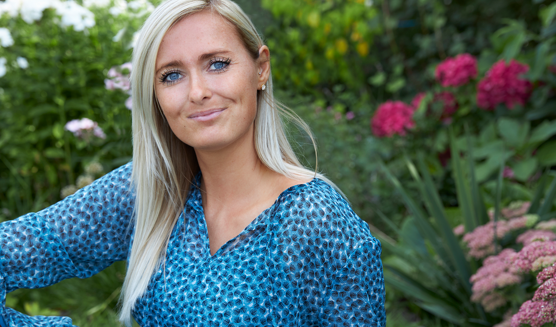 Sabines historie 3 Smilende kvinde med lyst hår sidder udendørs i en blomstrende have.